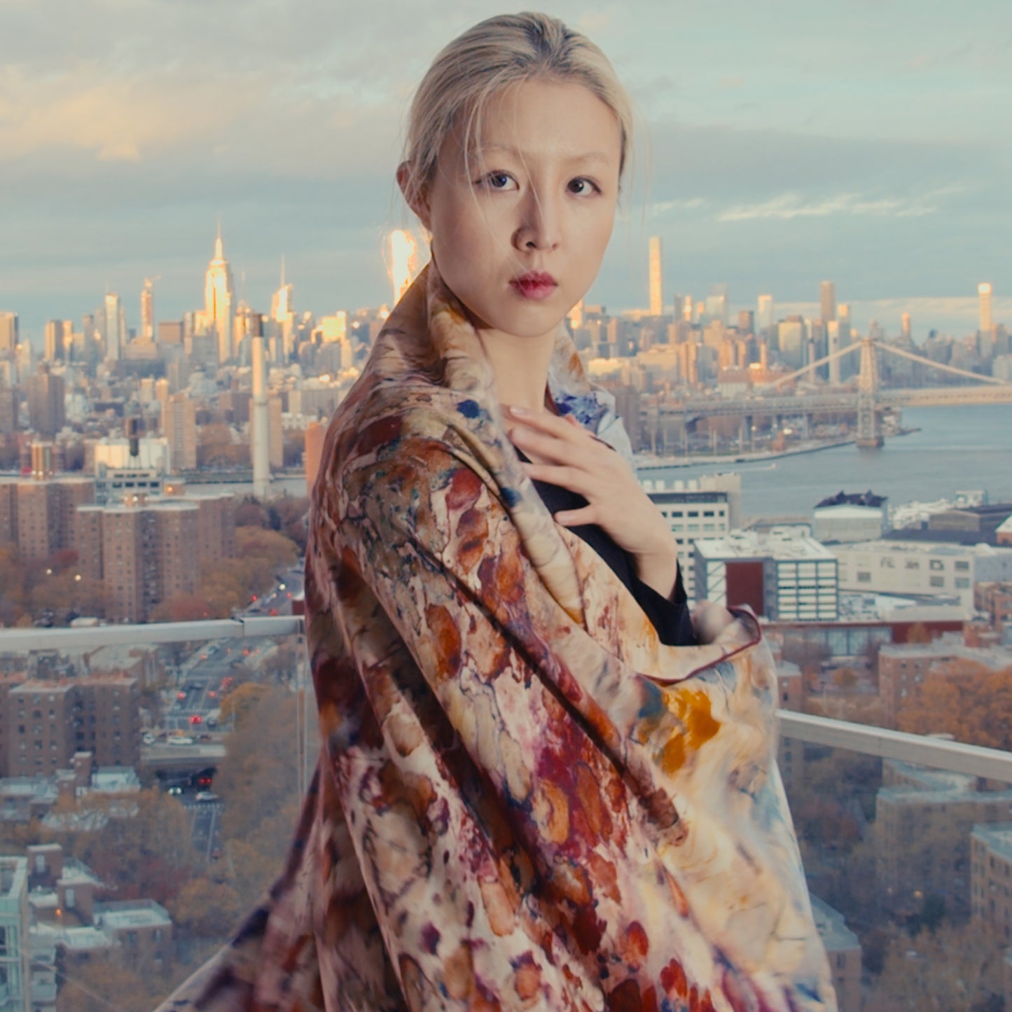 A model with bleach blonde hair elegantly wraps a vibrant scarf around herself, while the iconic Midtown Manhattan skyline serves as a stunning backdrop
