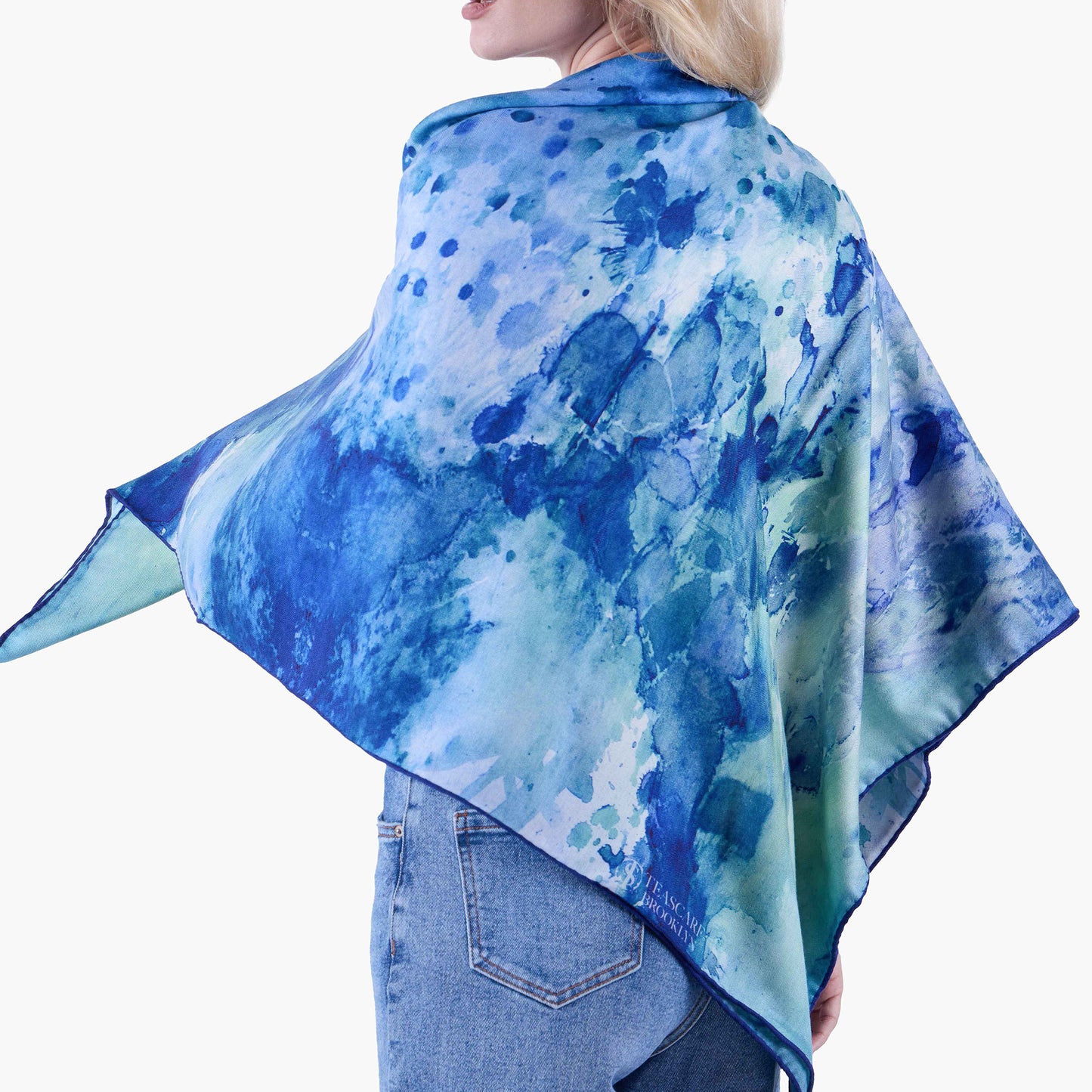 A back view of a model wearing an oversized silk-wool scarf in a soothing clear blue hue, elegantly draped to showcase its luxurious texture and lightweight design. The scarf features a subtle sheen, enhancing its rich, vibrant color. Perfect for versatile styling, it adds a touch of sophistication to any outfit.