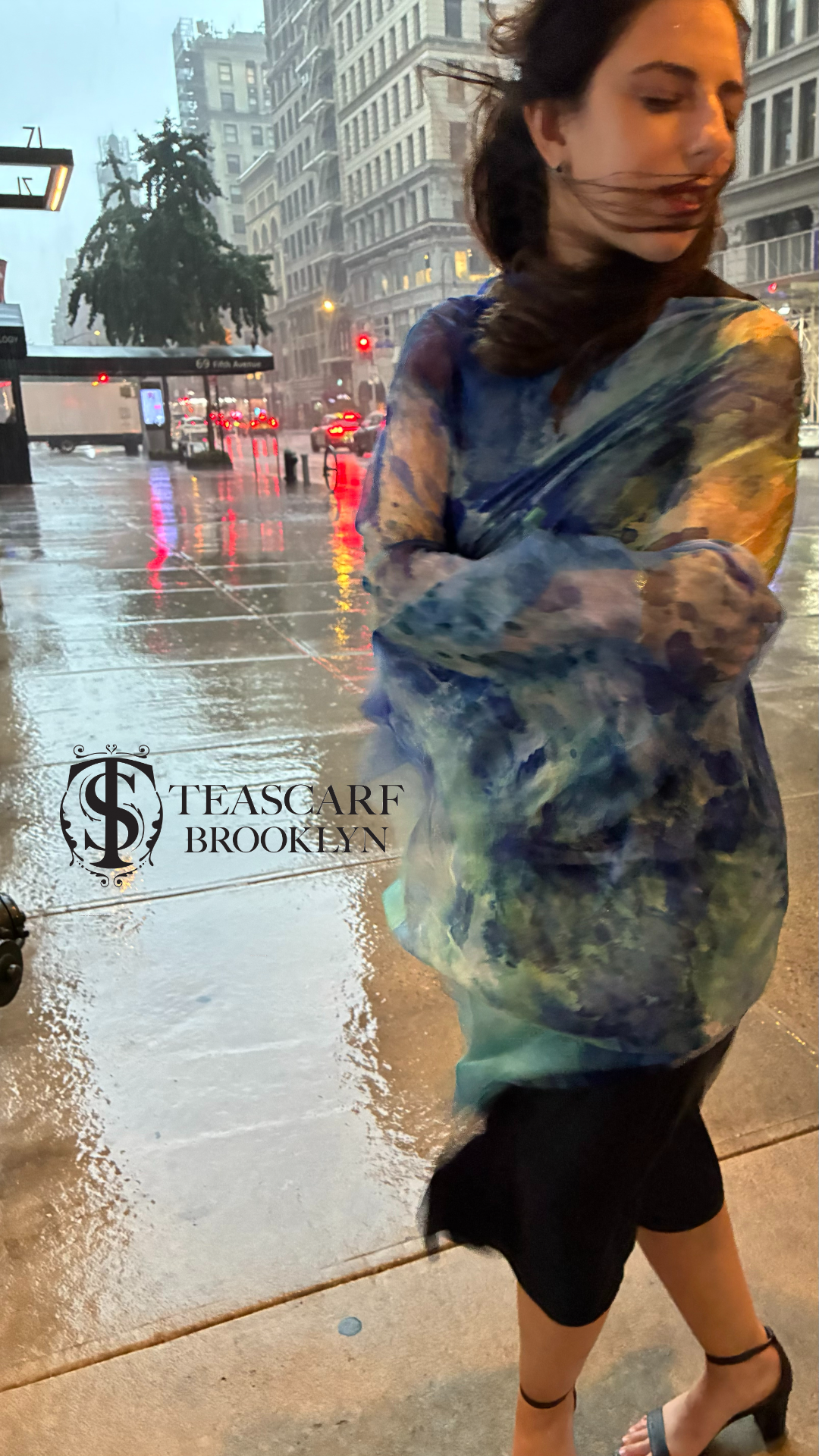 Woman wearing a Clear Blue scarf on a rainy, windy city street with 'TeaScarf Brooklyn' branding.