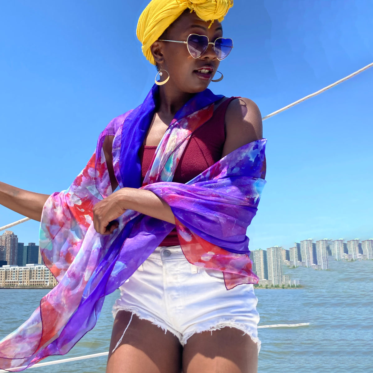 Model wears a purple rain, silk chiffon scarf over her shoulders and drapes a handkerchief around her neck while the breeze blows. on a sailboat.