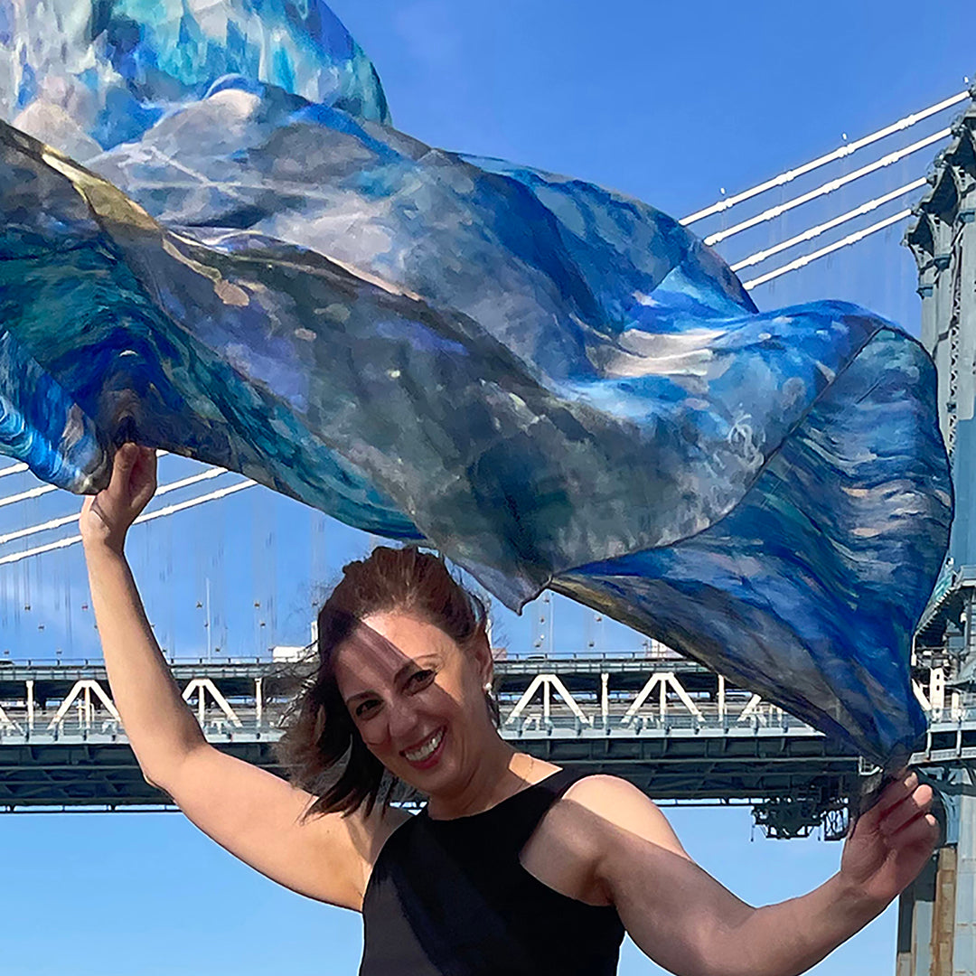 Model holding Blue Dream Oversized Square Tea-Printed Silk Chiffon Scarf billowing in the wind beneath a suspension bridge - 100% Silk, 52x52" - Teascarf Brooklyn