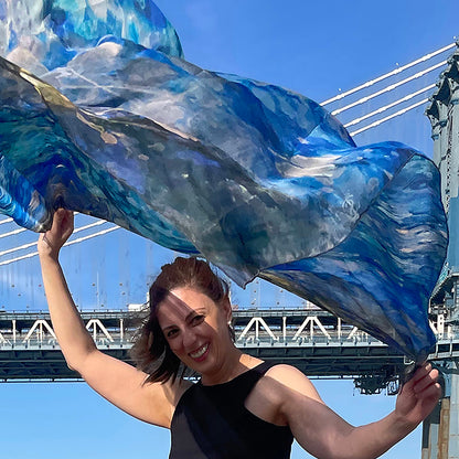 Model holding Blue Dream Oversized Square Tea-Printed Silk Chiffon Scarf billowing in the wind beneath a suspension bridge - 100% Silk, 52x52" - Teascarf Brooklyn