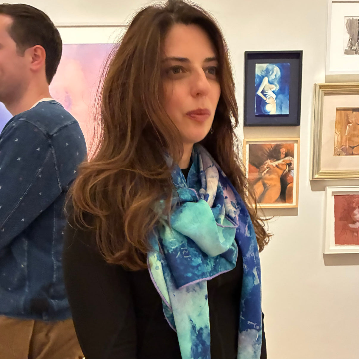 Woman wearing a colorful, 52x52" Peacock Green Oversized scarf in an art gallery setting with framed paintings on the wall. Wear art, then go see some.