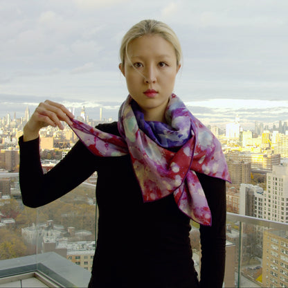 Video showcasing a vibrant silk-wool scarf from Teascarf Brooklyn, highlighting its luxurious texture, unique design, and rich colors as it gently drapes and flows in natural light. Model stands on 22nd floor view of Manhattan from Brooklyn.