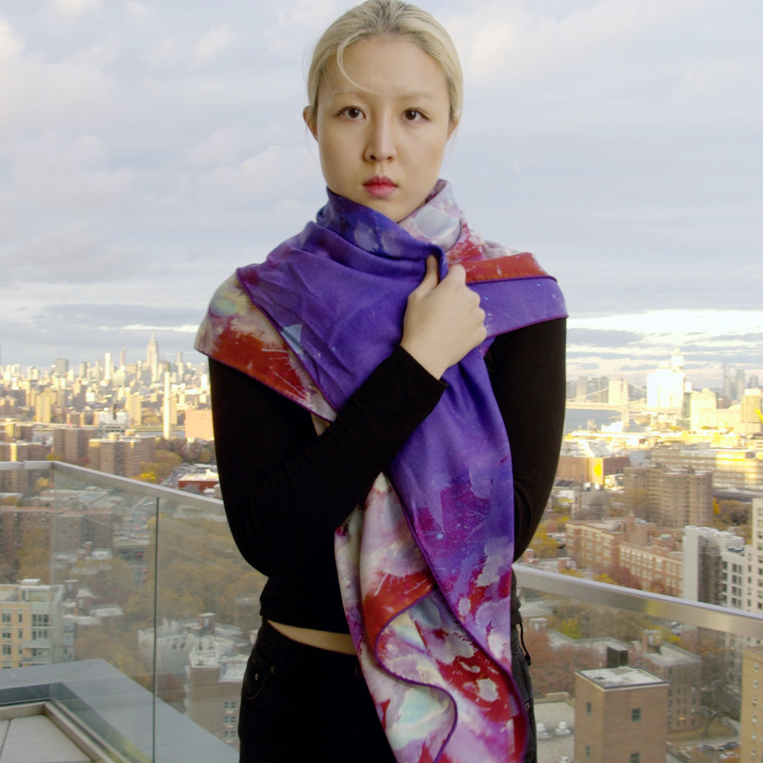 Video showcasing a vibrant silk-wool scarf from Teascarf Brooklyn, highlighting its luxurious texture, unique design, and rich colors as it gently drapes and flows in natural light. Model stands on 22nd floor view of Manhattan from Brooklyn.