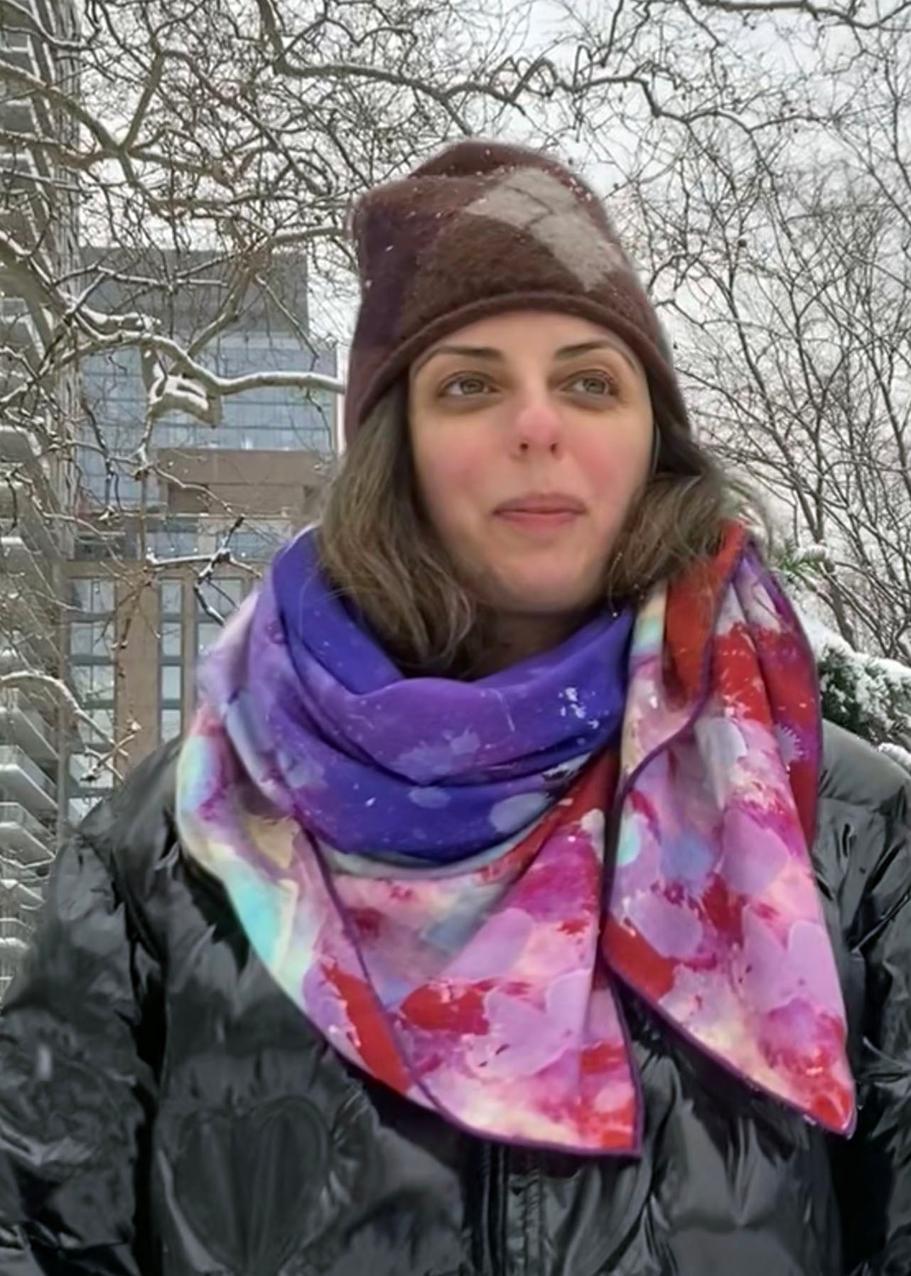 A woman stands outdoors in the snow, wearing a vibrant, tea-printed scarf in shades of purple, red, and white. She has a brown knit hat and a black puffer jacket, with snowflakes visible on her clothing. Bare trees and a modern building are in the background.