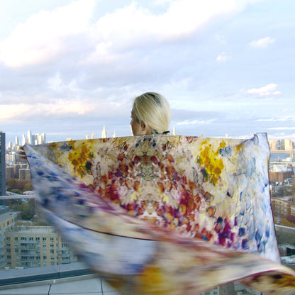 An elegant oversized scarf flutters in the breeze as model faces away made from a luxurious blend of silk and wool, featuring unique, abstract patterns made with steeped tea bags. The scarf showcases a mix of soft and earthy tones, with a lightweight yet warm fabric