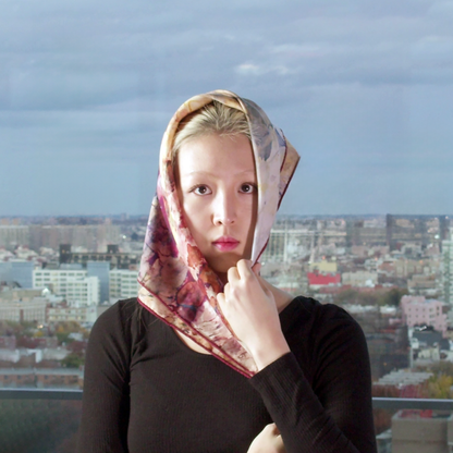 Model stands in front of city view, silk wool scarf is folded diagonally and being held on top of her head.