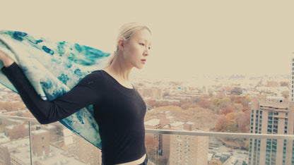 Video of a model on a roof overlooking Brooklyn wearing an oversized silk-wool scarf in a soothing clear blue hue, elegantly draped to showcase its luxurious texture and lightweight design. The scarf features a subtle sheen, enhancing its rich, vibrant color. Perfect for versatile styling, it adds a touch of sophistication to any outfit.